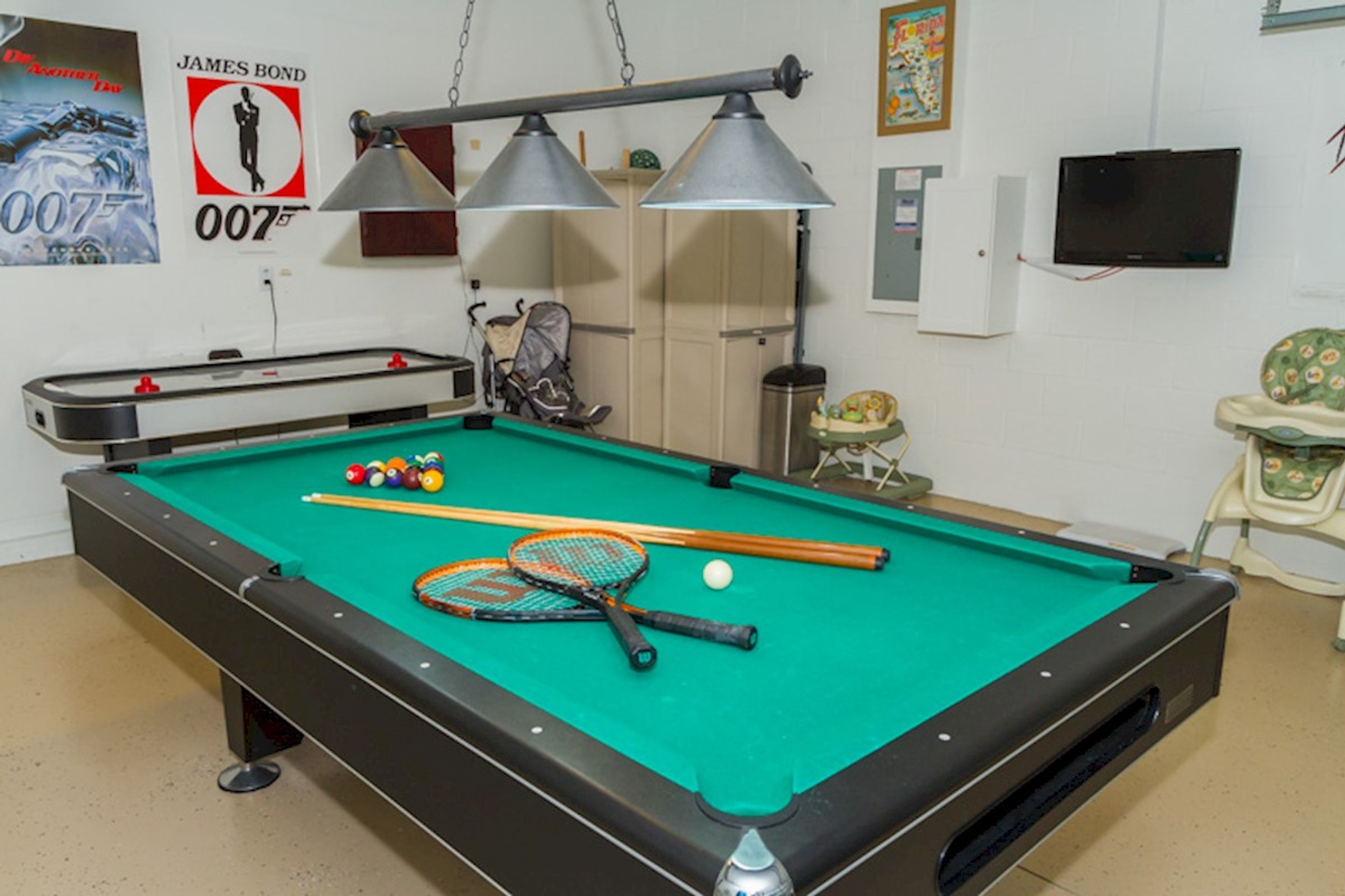 A pool table with cues, a pool ball, and a table tennis paddle. There is a baby stroller and a television mounted on the wall.