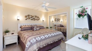 Une chambre à coucher comprenant un lit, des tables de chevet, un ventilateur de plafond, des appliques murales, un miroir et une fenêtre avec des stores.