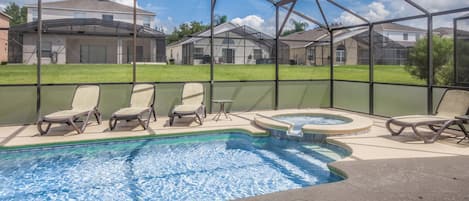A swimming pool with lounge chairs and a hot tub, surrounded by a screened area with houses in the background.