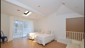 Un dormitorio con dos camas, un ventanal con cortinas, un tragaluz y una barandilla blanca.