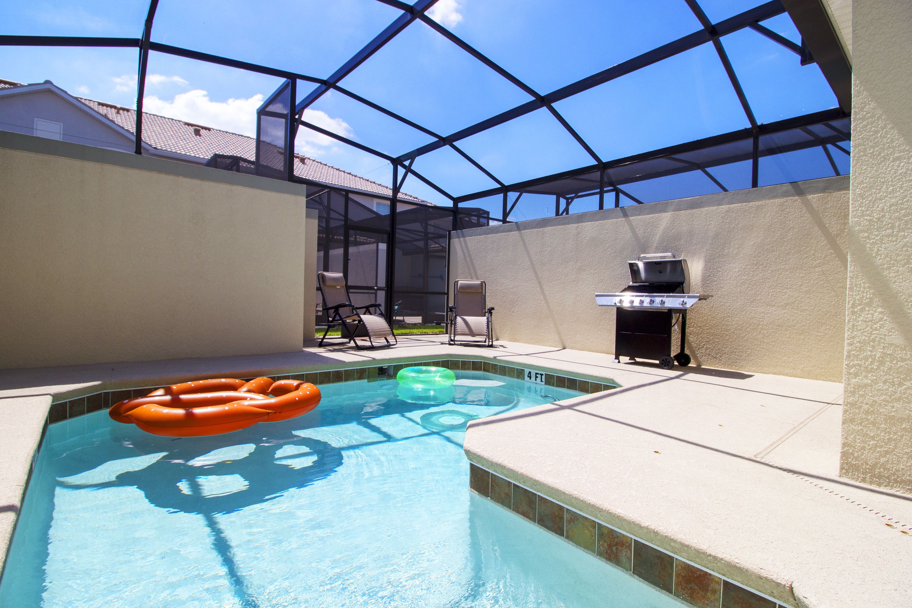 A swimming pool with a glass enclosure, a small grill, and lounge chairs.