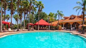 Una piscina con sillones de descanso y toldos rojos, rodeada de palmeras y un edificio con techo de tejas.