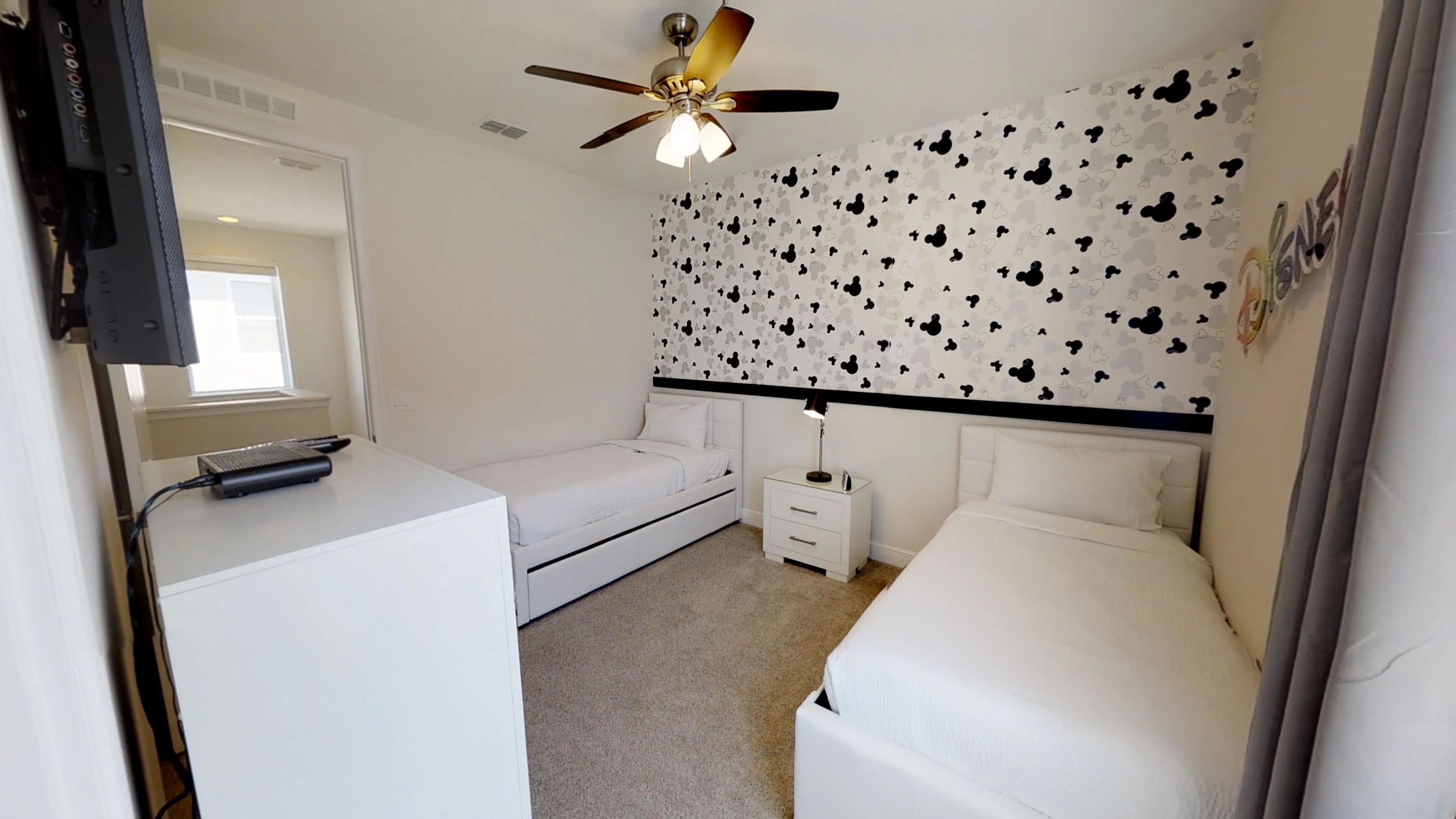 Une chambre avec deux lits, un bureau, un ventilateur de plafond et un mur décoré d’un motif de Mickey Mouse.