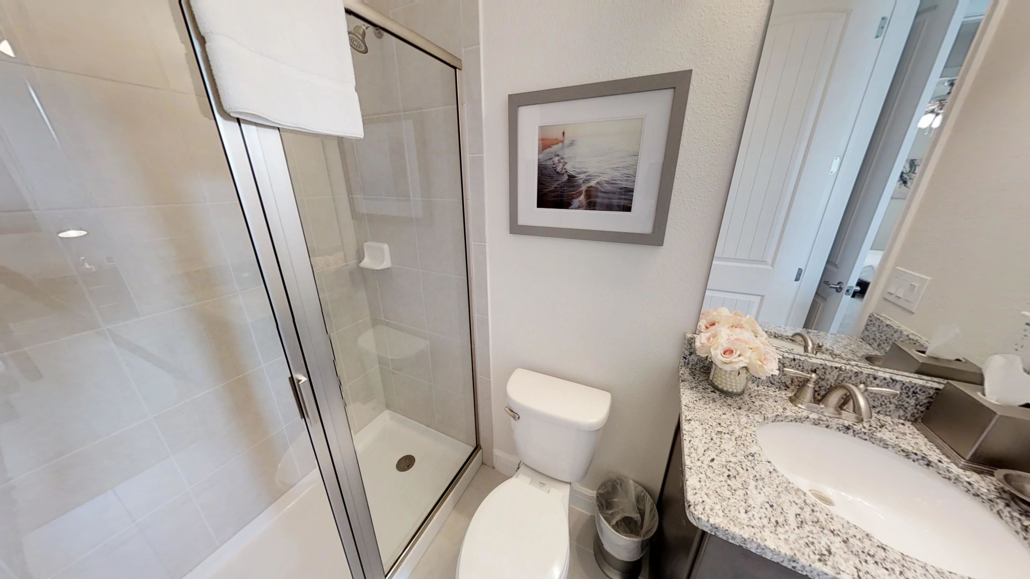 A modern bathroom with a glass shower enclosure, a toilet, a sink with a granite countertop, and a framed picture on the wall.