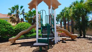 A playground with climbing structures and slides, surrounded by palm trees and a building in the background. - Regal Palms Resort & Spa 1215 (Davenport)