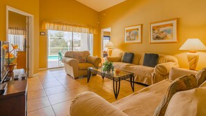 A living room with two sofas, a coffee table, and a TV. There is a window with curtains and a door leading outside.