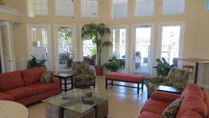 A spacious living room with a red sofa, a glass coffee table, and patterned armchairs. There are large windows allowing natural light and a view of the outdoors.