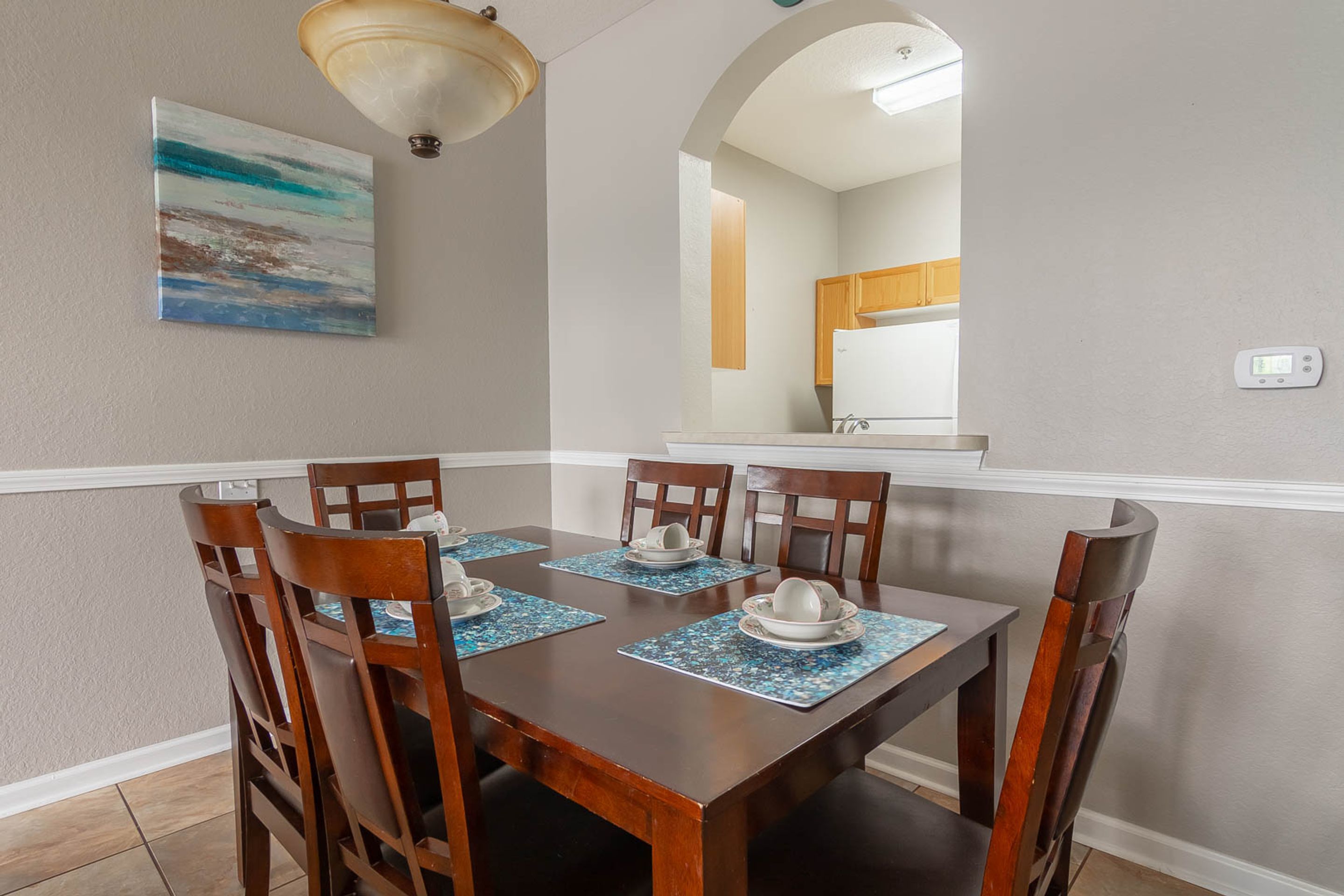 Uma sala de jantar com uma mesa de madeira posta para seis pessoas, seis cadeiras e um quadro na parede.