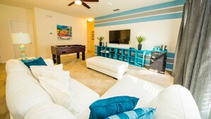 A living room with a sofa, ottoman, and a foosball table. There is a TV mounted on the wall and a ceiling fan. - Solterra 4043 (Davenport)