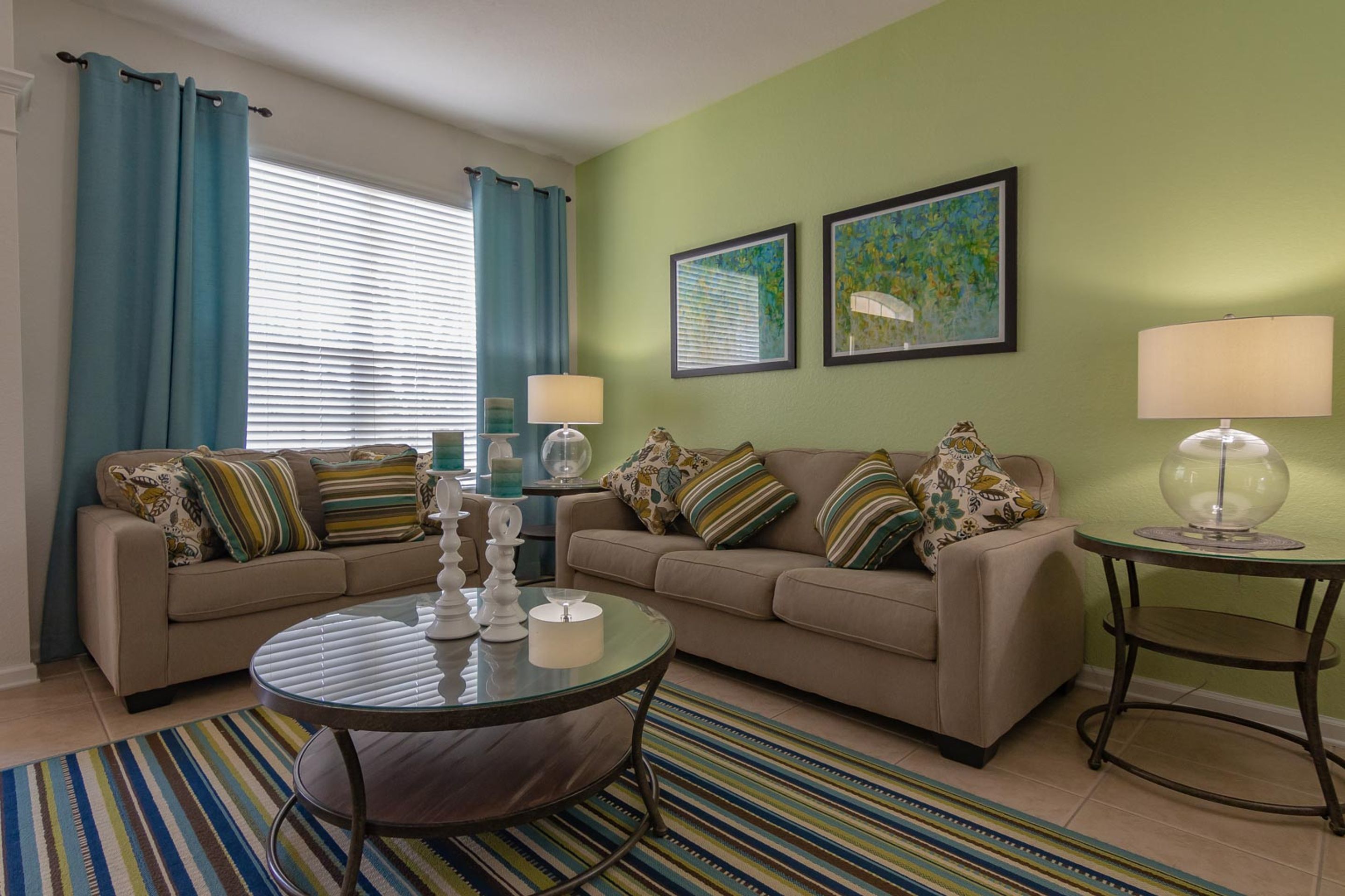 Una sala de estar con un sofá color beige, dos mesitas auxiliares, una mesa de centro de vidrio, un tapete a rayas y una ventana con cortinas azules.
