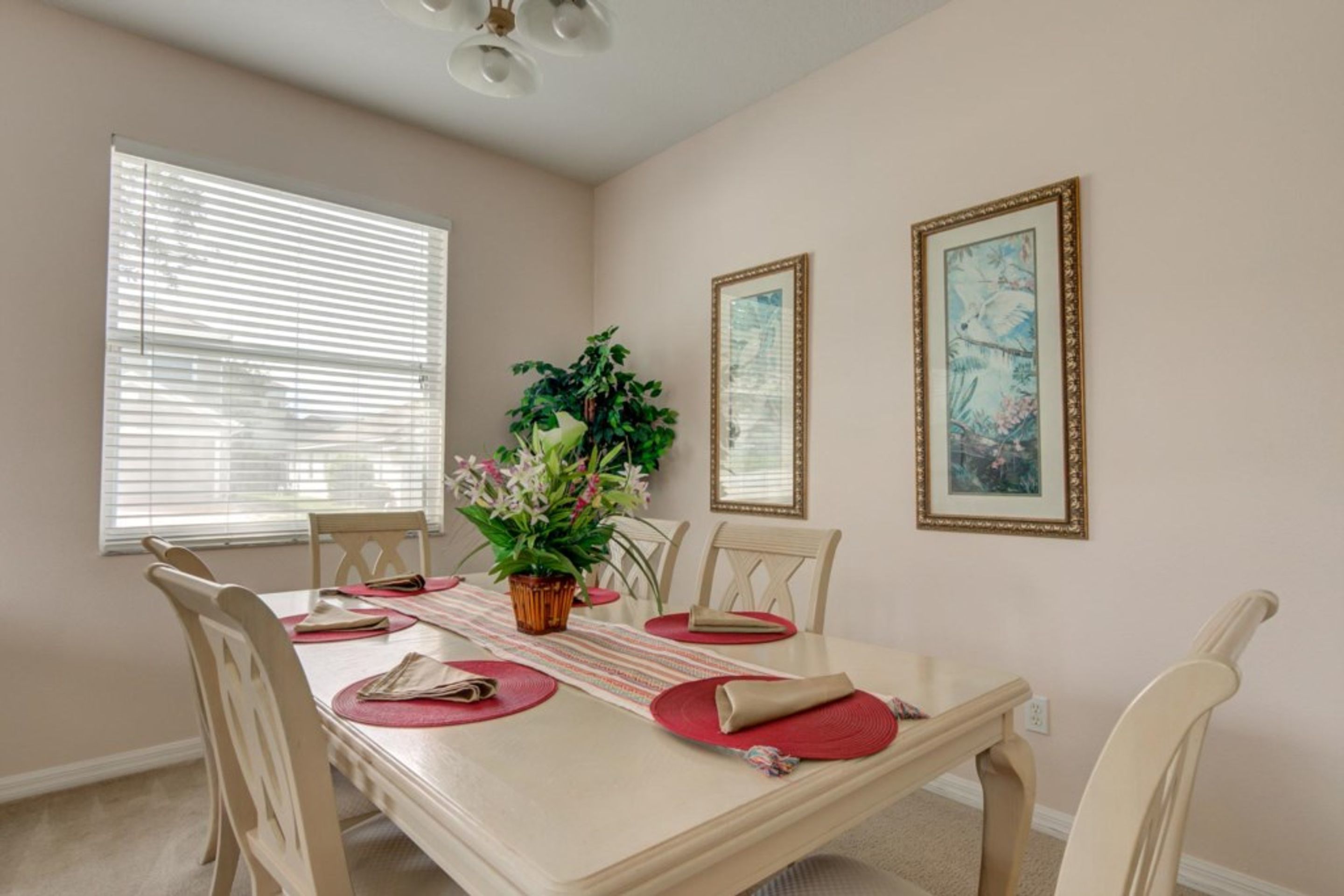 A dining room with a table set for four, surrounded by chairs, a potted plant, and two framed artworks on the wall.