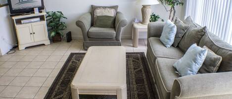A living room with a sofa, armchair, coffee table, TV, lamp, and plants.