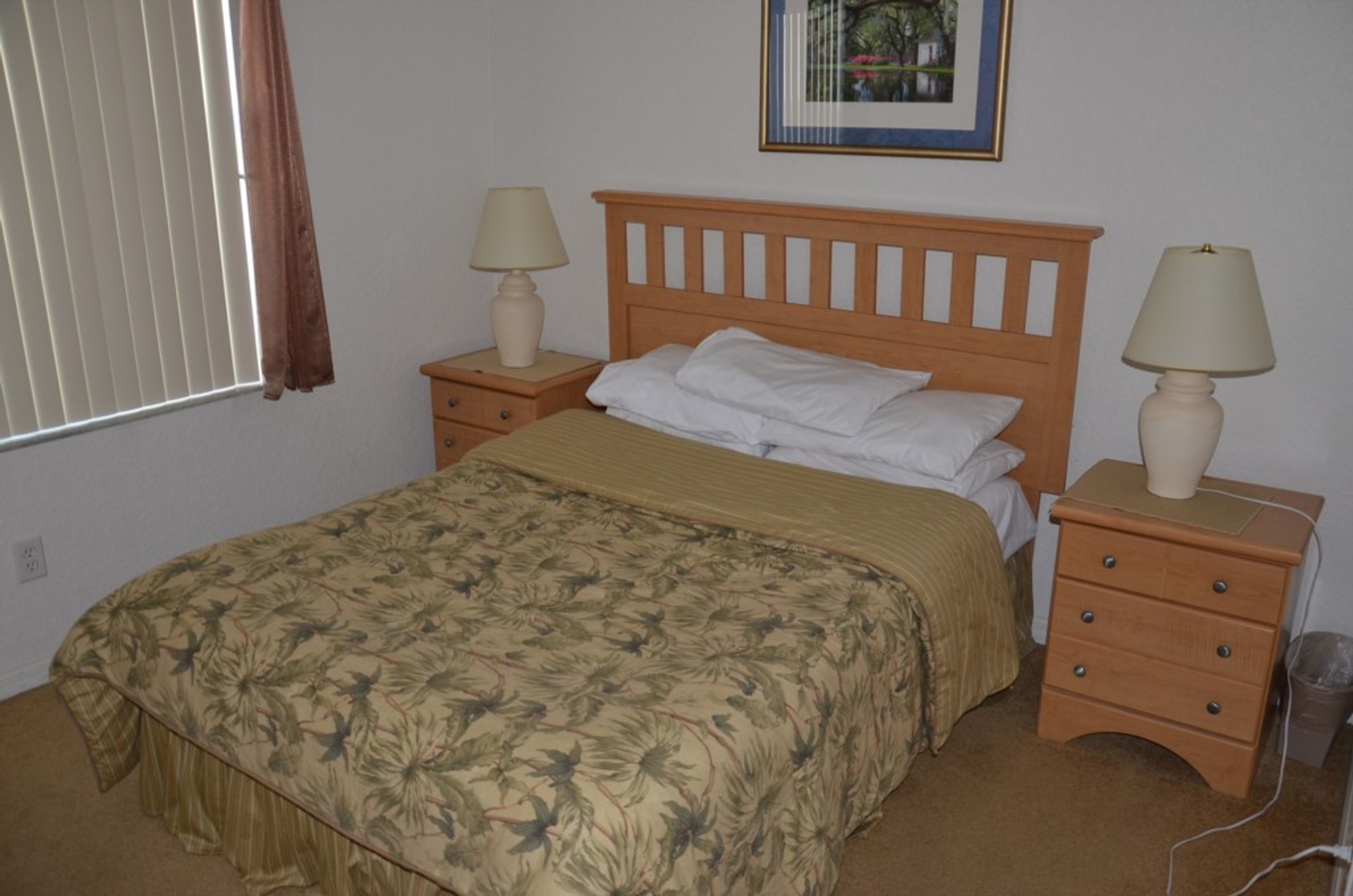 A bedroom with a bed, two bedside tables with lamps, a window with blinds, and a framed picture on the wall.
