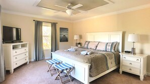 A bedroom with a bed, two bedside tables, a TV, a ceiling fan, a window with curtains, and a small stool.