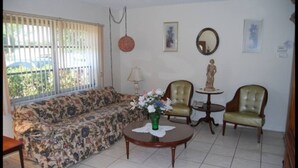 A living room with a floral sofa, two green armchairs, a round coffee table with a vase of flowers, a small table with a statue, and a window with blinds. - Bonita Springs 23 (Bonita Springs)