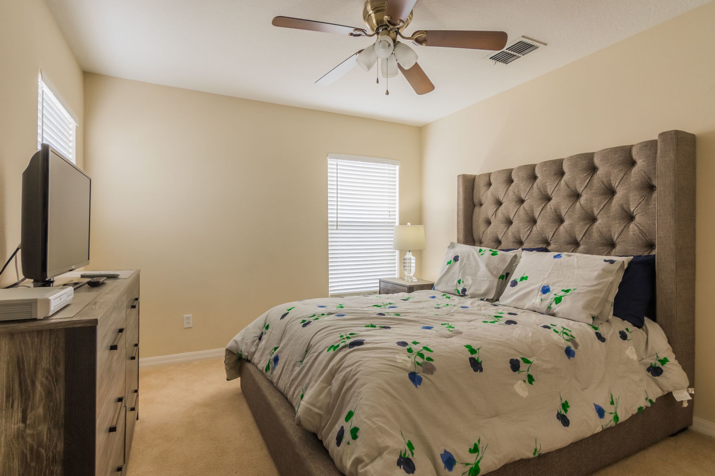 Une chambre à coucher avec une tête de lit capitonnée, un lit recouvert d’une literie à motifs floraux, un ventilateur de plafond, un téléviseur posé sur une commode en bois et une fenêtre avec des stores.