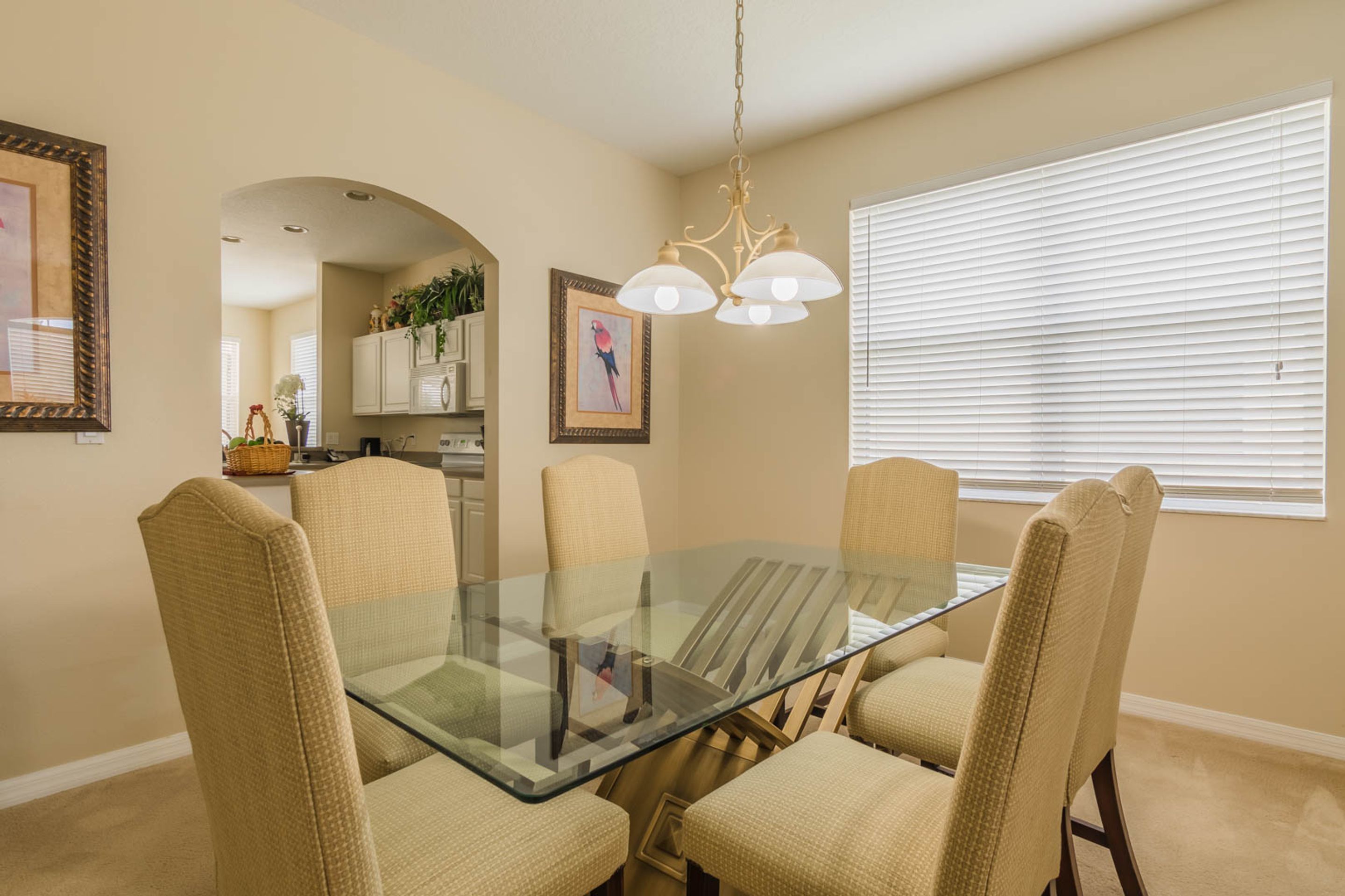 A dining area with a glass-top table surrounded by six beige chairs, a chandelier, and a window with blinds.