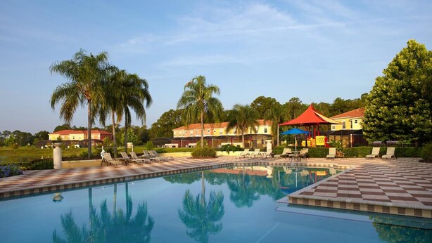 A swimming pool with lounge chairs and umbrellas, surrounded by palm trees and buildings. - Encantada Resort 3076 (Kissimmee)