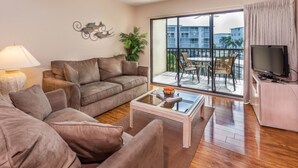 A living room with a sofa, coffee table, and a TV. There is a balcony with a table and chairs. - Fort Myers Beach Area 7327 - 203 (Fort Myers Beach)