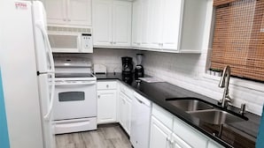 A kitchen with white cabinets, a black countertop, a stainless steel sink, a refrigerator, a stove, and a coffee maker. - Fort Myers Beach Area 7327 - 203 (Fort Myers Beach)