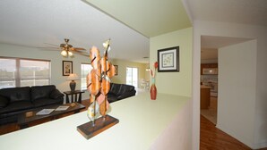 A living room with a black leather sofa, a coffee table, and a decorative sculpture. There is a ceiling fan, a framed picture on the wall, and a kitchen in the background. - Four Corners 908 (Davenport)