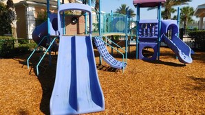 A playground with slides and a climbing structure, surrounded by palm trees and a building in the background. - Coral Cay 2475 (Kissimmee)