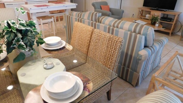 A living room with a sofa, a glass coffee table, and a dining table with plates and bowls. There is a kitchen area with a microwave and stove in the background. - Coral Cay 2475 (Kissimmee)