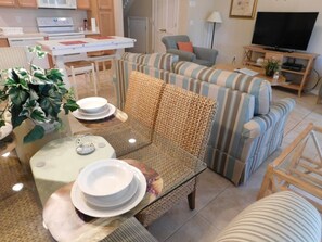 A living room with a sofa, a glass coffee table, and a dining table with plates and bowls. There is a kitchen area with a microwave and stove in the background. - Coral Cay 2475 (Kissimmee)