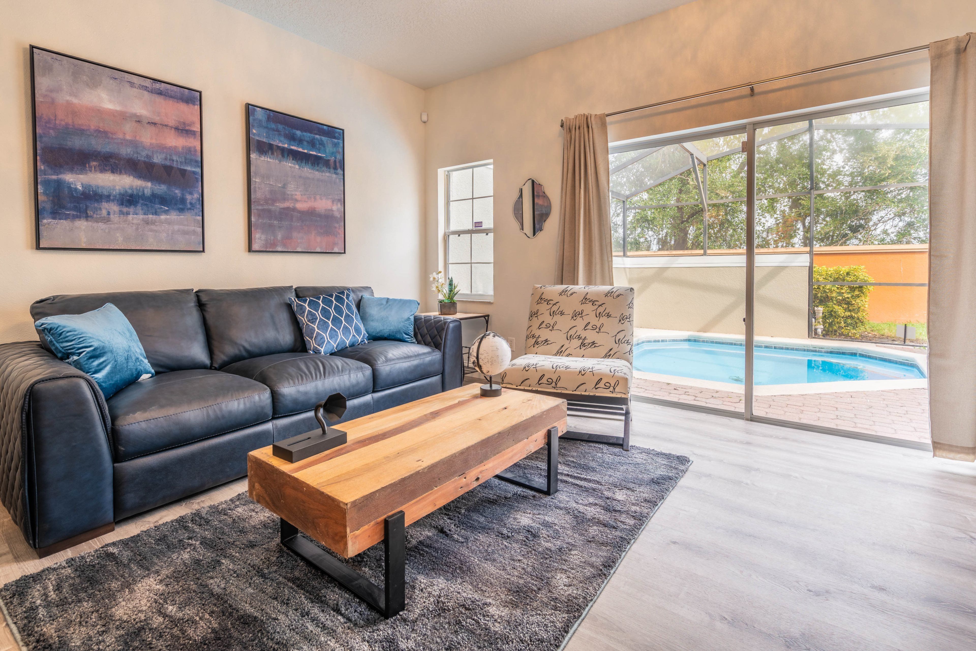 A living room with a black leather sofa, a wooden coffee table, and a patterned armchair. A large window offers a view of a swimming pool.