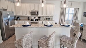 A kitchen with white cabinets, stainless steel appliances, a central island with bar stools, and pendant lighting. - Golden Palms Vacation Resort 2588 (Kissimmee)