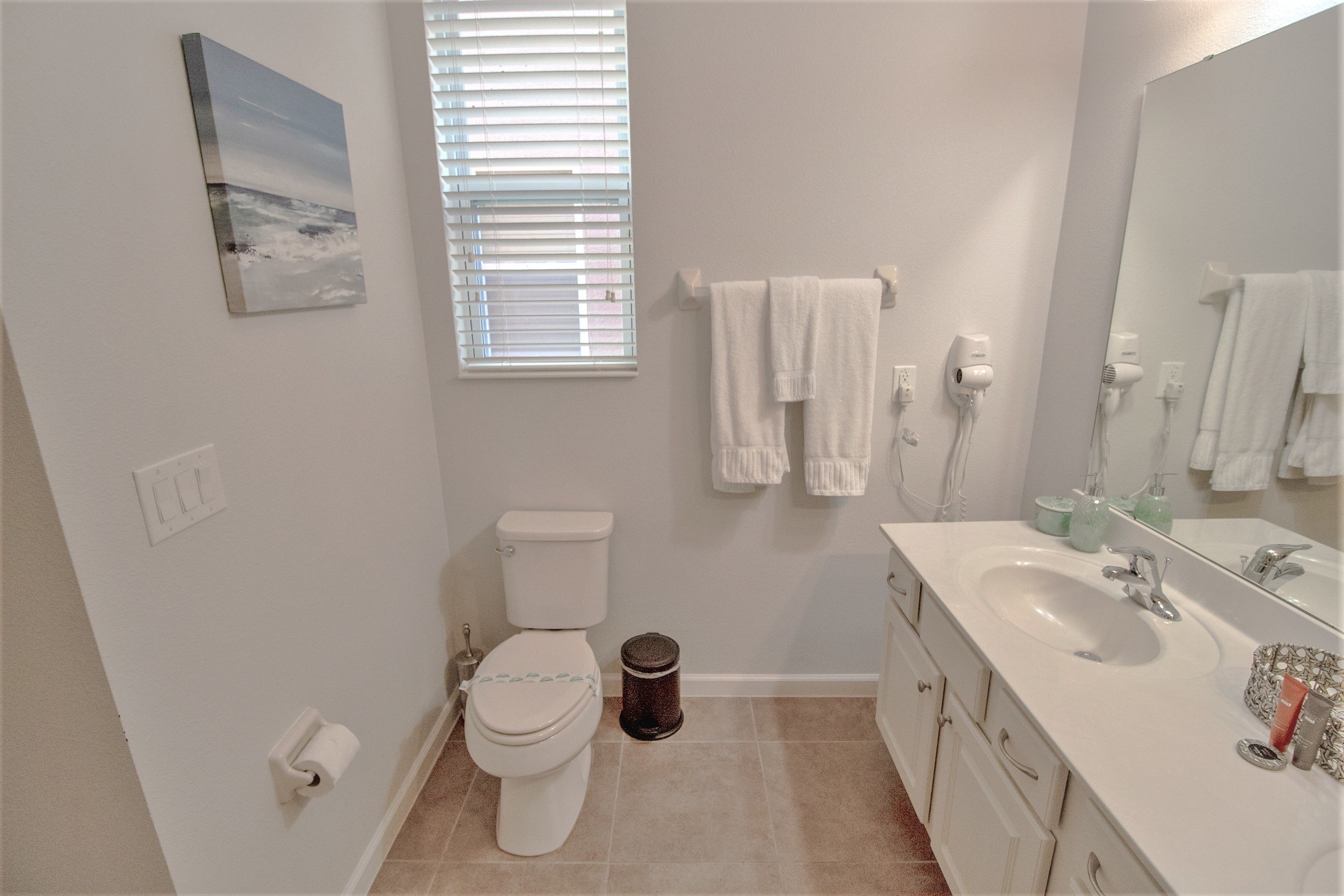 A bathroom with a toilet, sink, and mirror. There is a window with blinds, a towel rack, and a trash can.