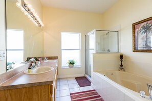 A bathroom with two sinks, a shower, and a bathtub. There is a window with a plant on the windowsill, and a mirror with lights above the sink.