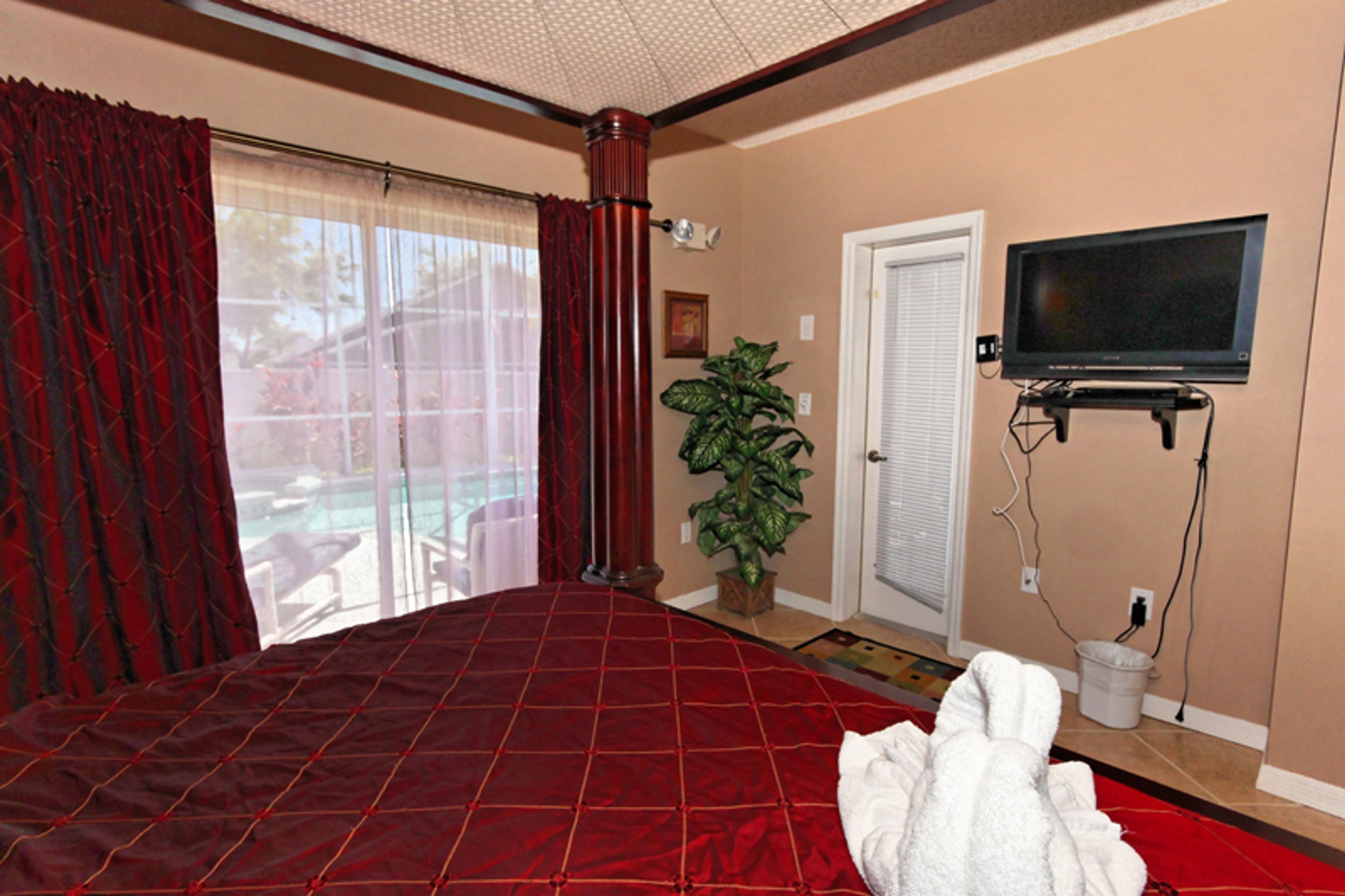 A hotel room with a large bed, a television mounted on the wall, a potted plant, and a door with a window.