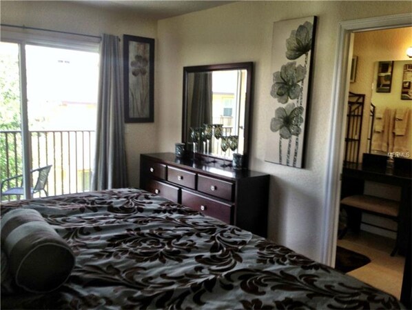 A bedroom with a bed, dresser, and a large mirror. There is a balcony with a chair and curtains.