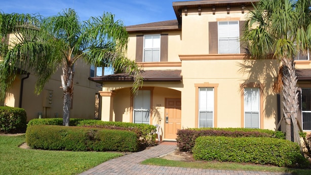 A two-story building with a beige facade, a brick pathway leading to the entrance, and well-maintained landscaping including hedges and palm trees.