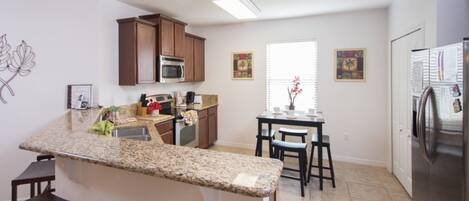 A kitchen with granite countertops, wooden cabinets, a microwave, and a refrigerator. There is a dining area with a table and chairs.