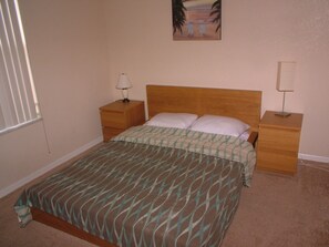 Quarto com cama, duas mesas de cabeceira, uma luminária em cada e um quadro sobre a cama, além de uma janela com persianas.