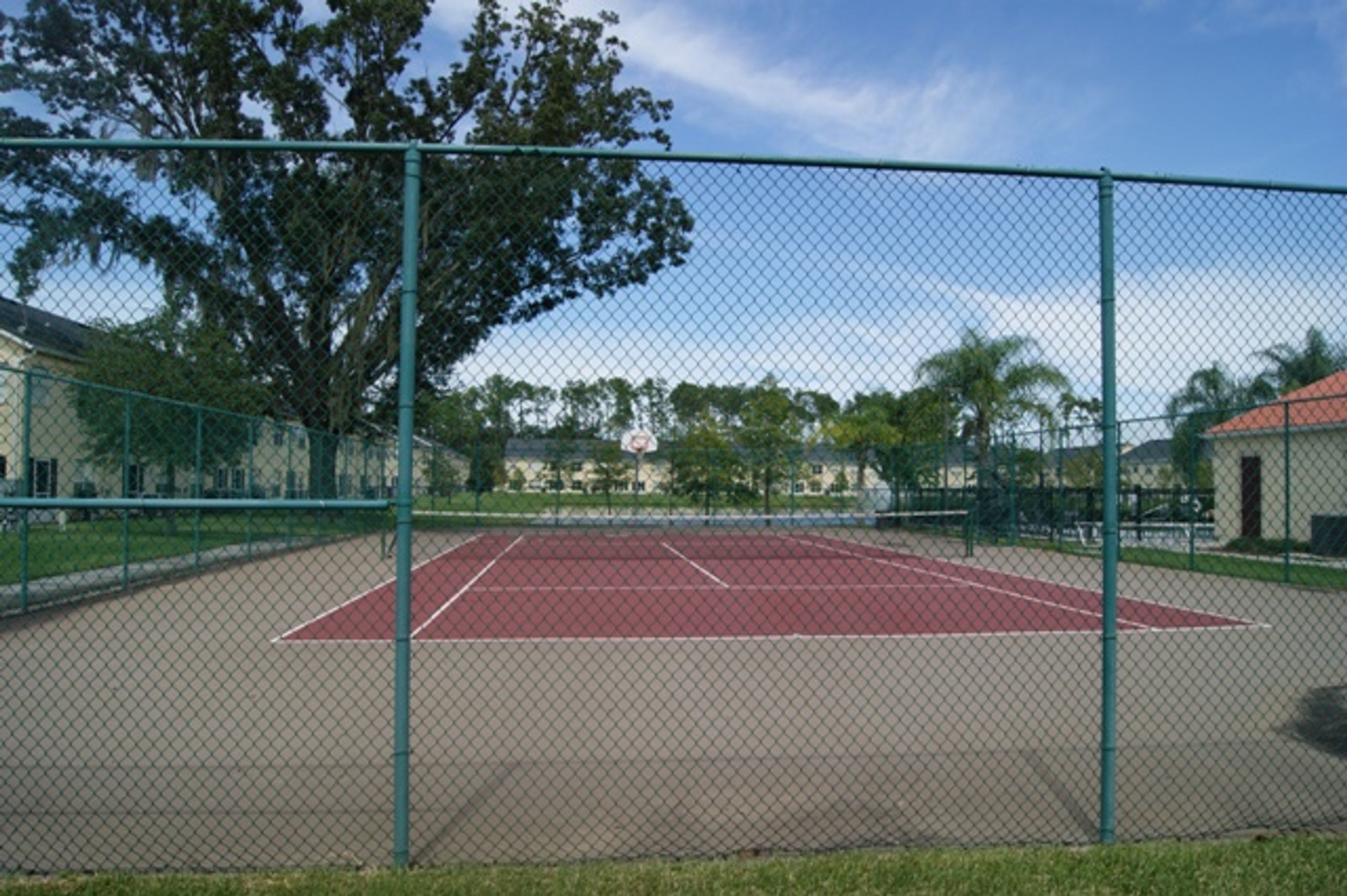 A tennis court enclosed by a chain-link fence, surrounded by trees and buildings.