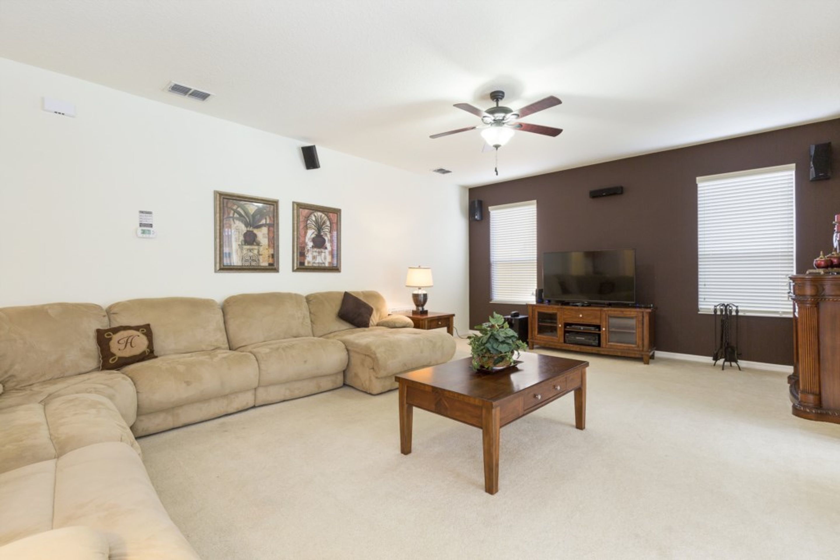 A living room with a beige sectional sofa, a wooden coffee table, a ceiling fan, a TV on a wooden stand, and a window with blinds.