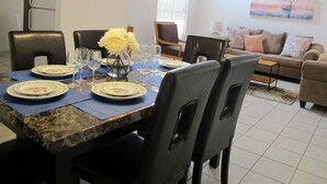 Uma área de refeições com mesa de mármore, cadeiras pretas e um vaso de flores amarelas. A mesa está posta com pratos, copos e um arranjo central.
