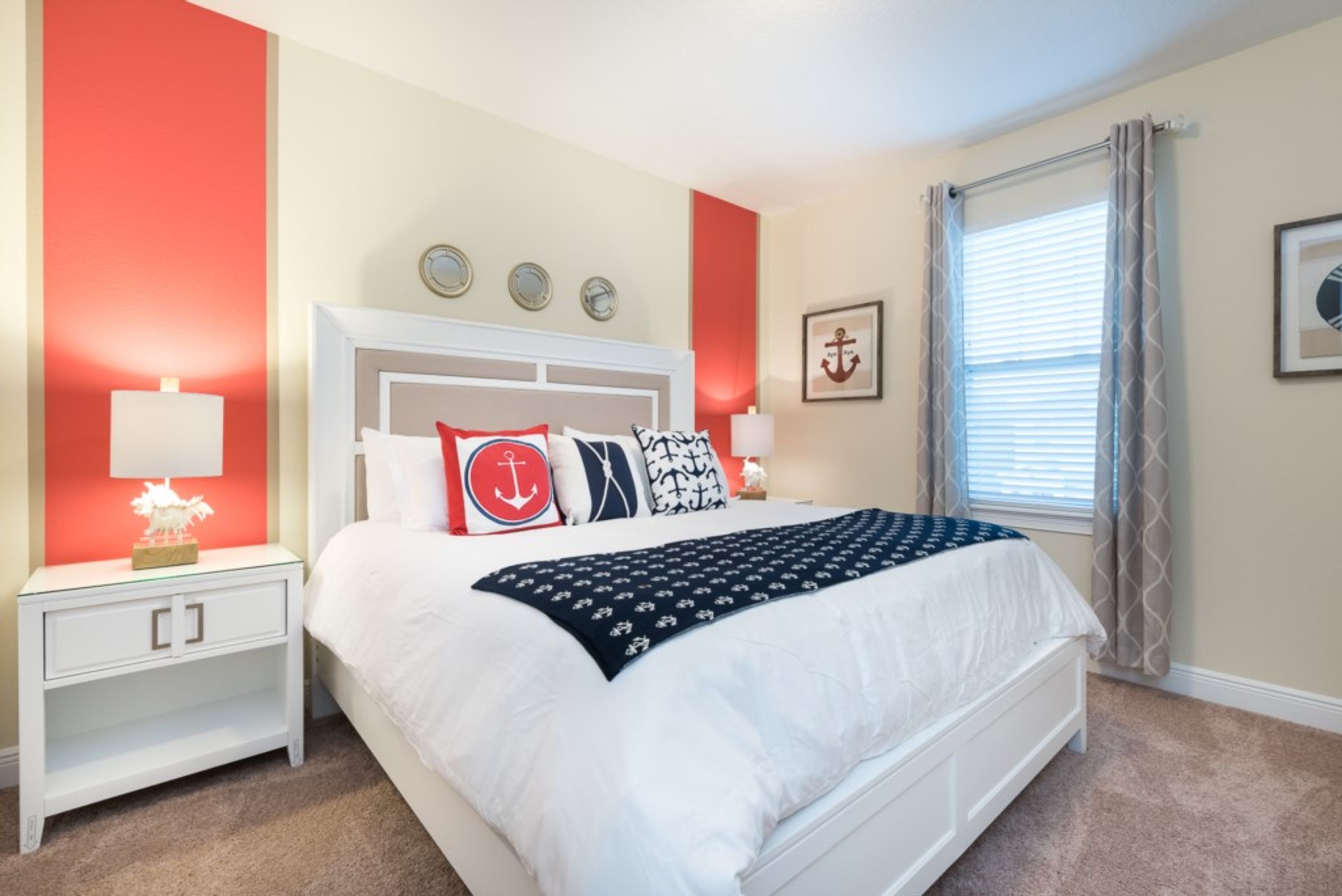 A bedroom with a white bed, red accent walls, a nightstand with a lamp, and a window with blinds.