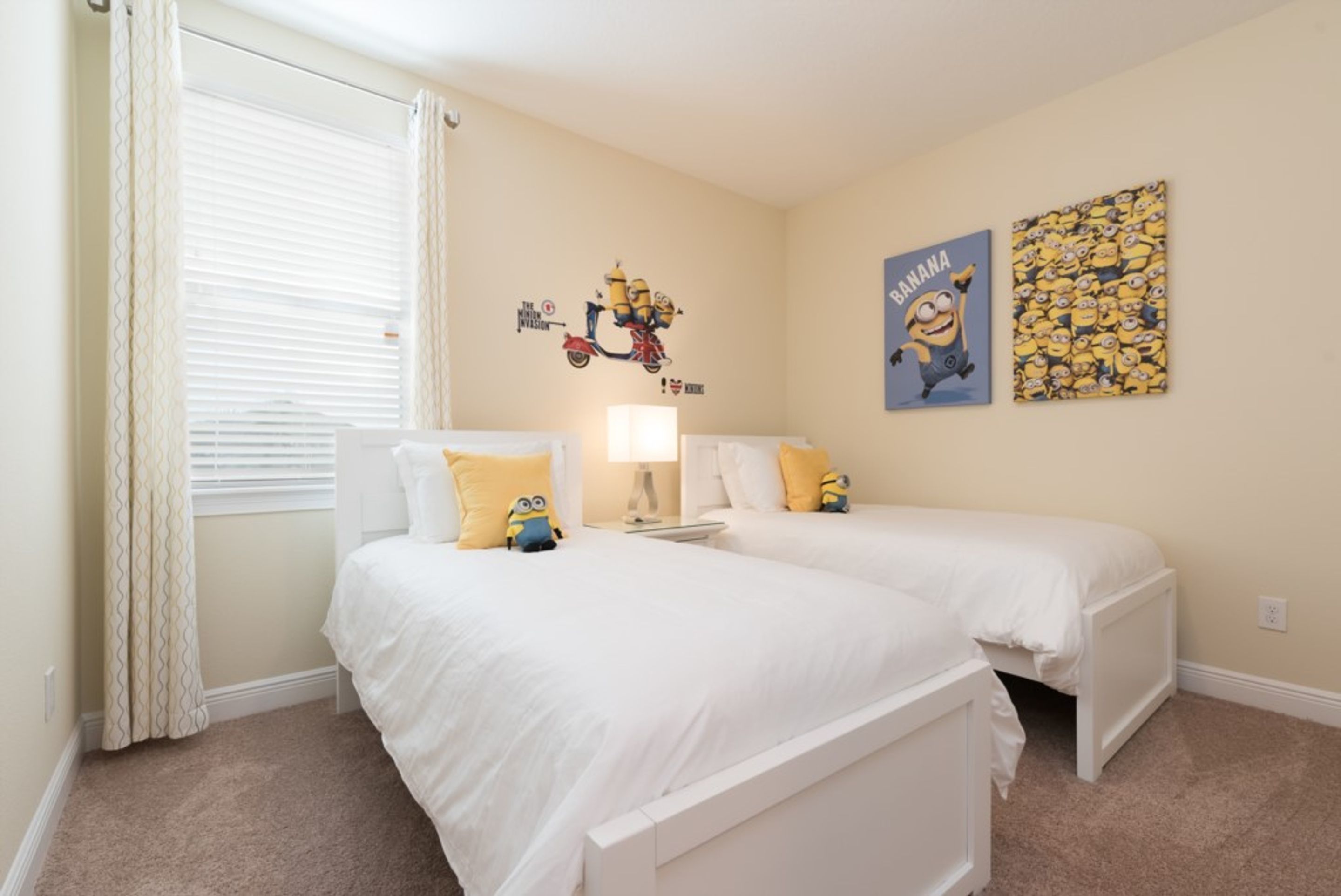 A bedroom with two beds, a window with blinds, and Minion-themed wall art.
