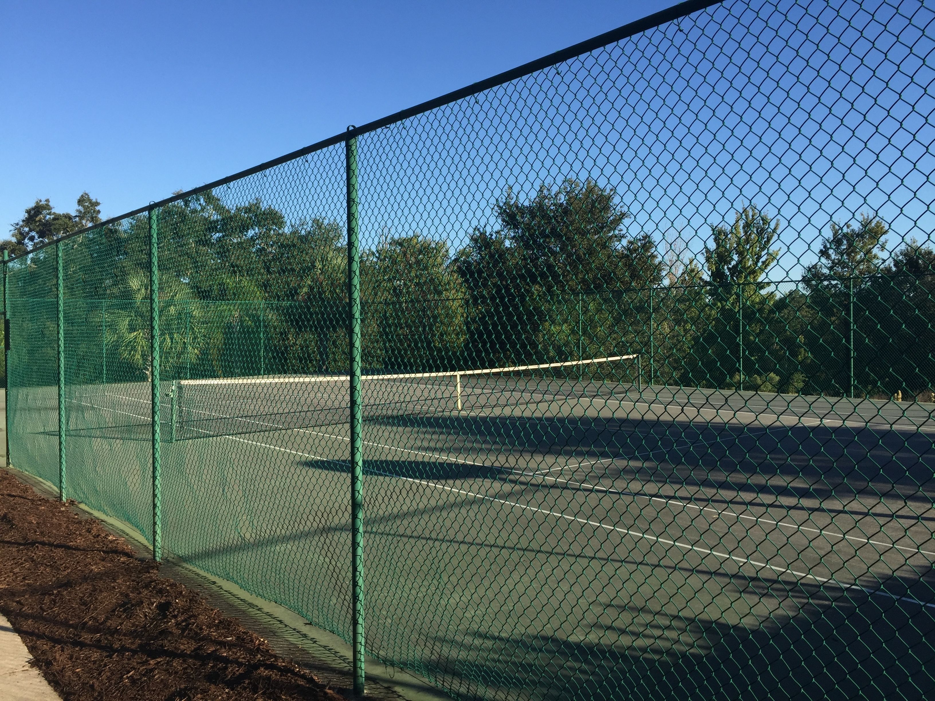 En tennisbane omgivet af et grønt hegn af pigtråd, med træer i baggrunden.