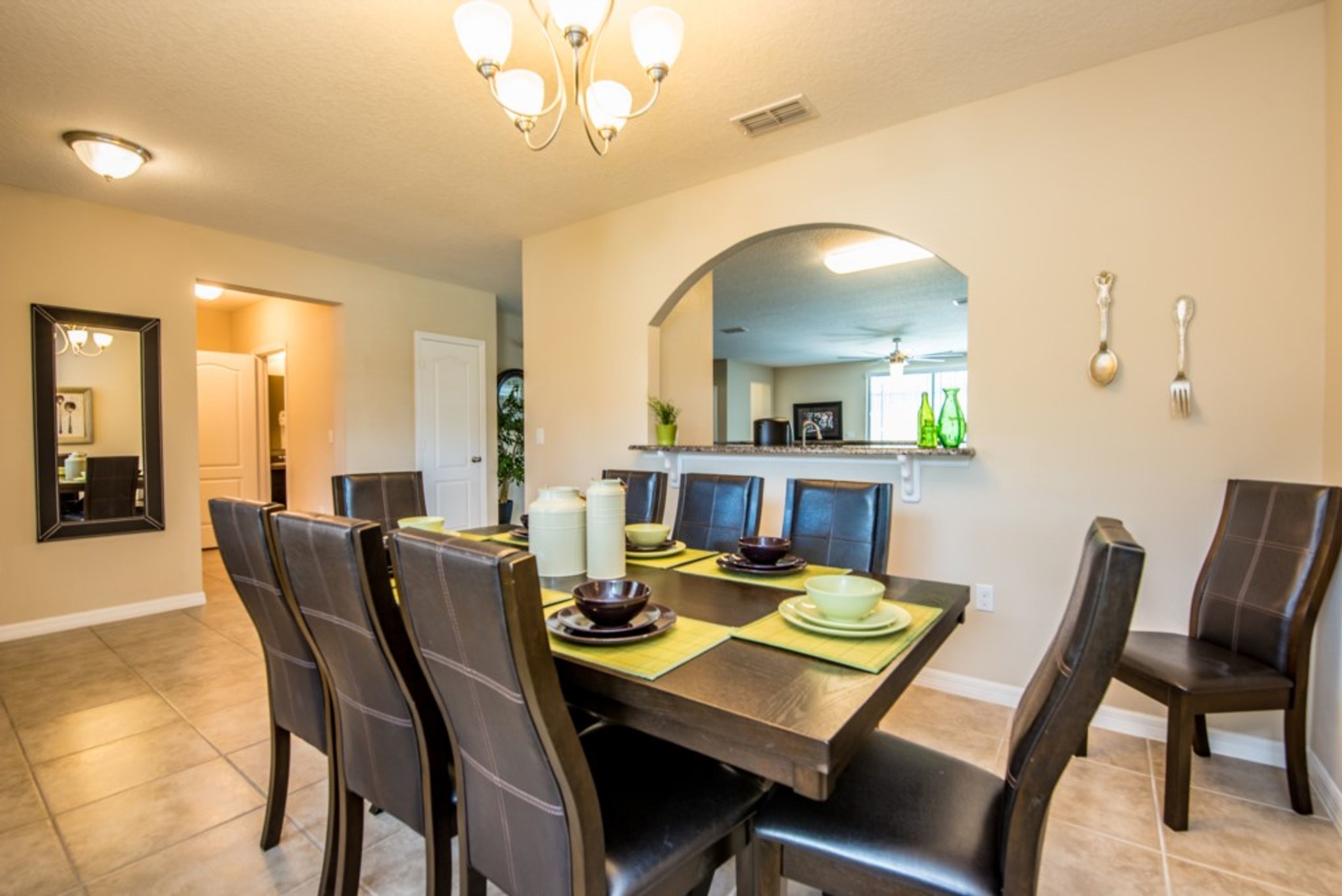A dining area with a large table, chairs, and a chandelier. There is a kitchen visible in the background.