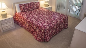 Un dormitorio con una colcha estampada, dos lámparas de noche, una mesita de noche y vistas a través de una ventana con persianas.