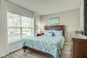 A bedroom with a large window, a bed with patterned bedding, a nightstand, a dresser, and a painting on the wall.