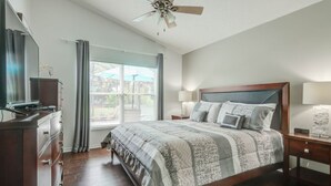 Une chambre à coucher comprenant un lit, une commode, une table de chevet, un ventilateur de plafond et une fenêtre avec des rideaux.