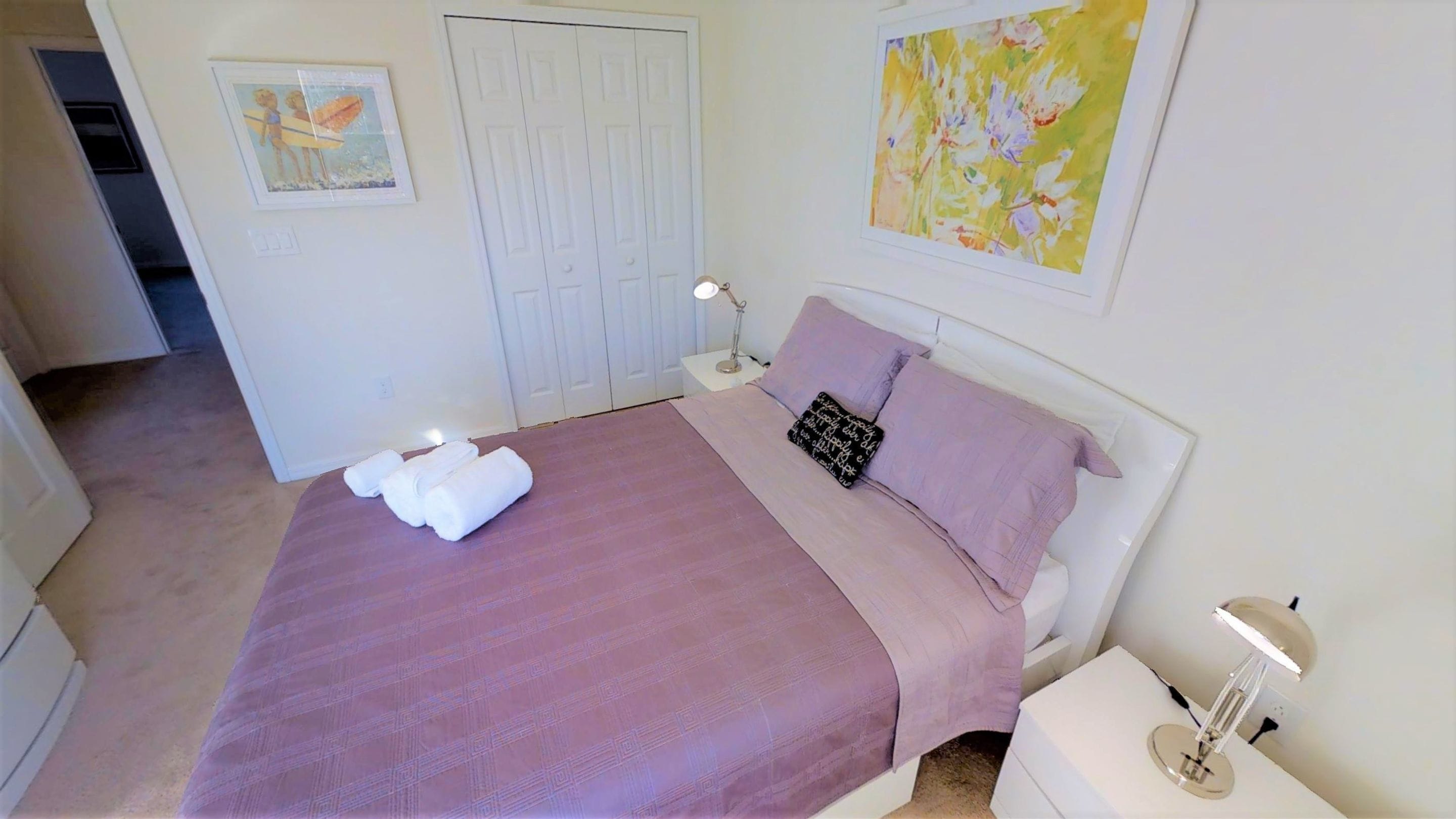 A bedroom with a bed, a nightstand, a lamp, a door, and a painting on the wall.