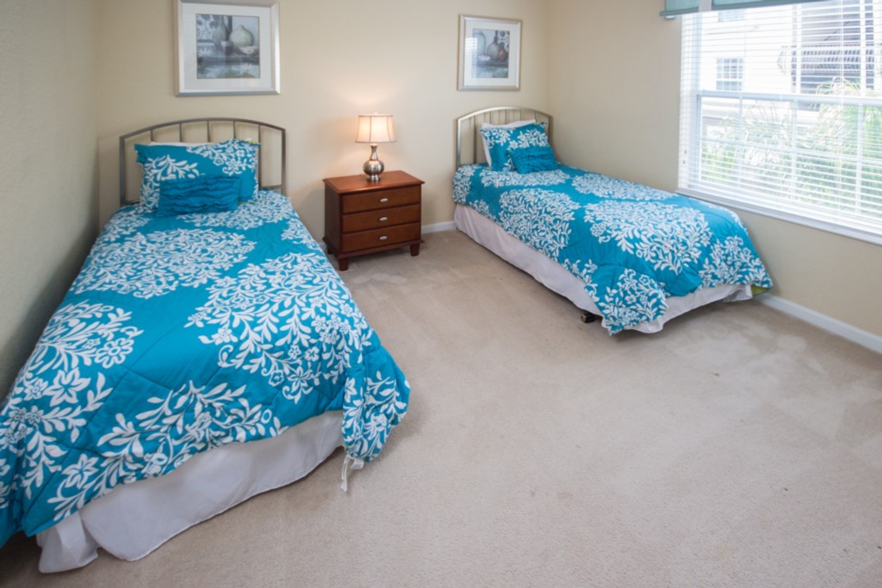 A bedroom with two beds, each with a blue floral bedspread and matching pillows. There is a nightstand with a lamp between the beds.