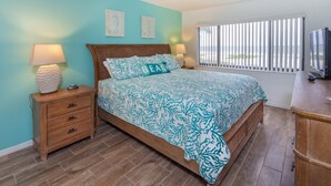 A bedroom with a bed, two bedside lamps, a wooden nightstand, a dresser, and a window with blinds.